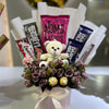 Bouquet with flowers, a teddy bear, and various chocolate bars on a white surface.