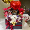 Gourmet gift basket with candy, flowers, and a teddy bear on a white surface.