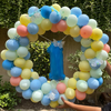 Balloon arch with pastel colors and decorative ribbons against a green hedge background