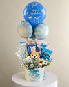 Pastel blue birthday gift basket with a teddy bear, snacks, chocolates, floral decorations and blue balloons.