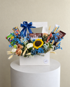 White gift box with a blue bow, chocolates, snacks, mixed floral decorations and a crochet sunflower.