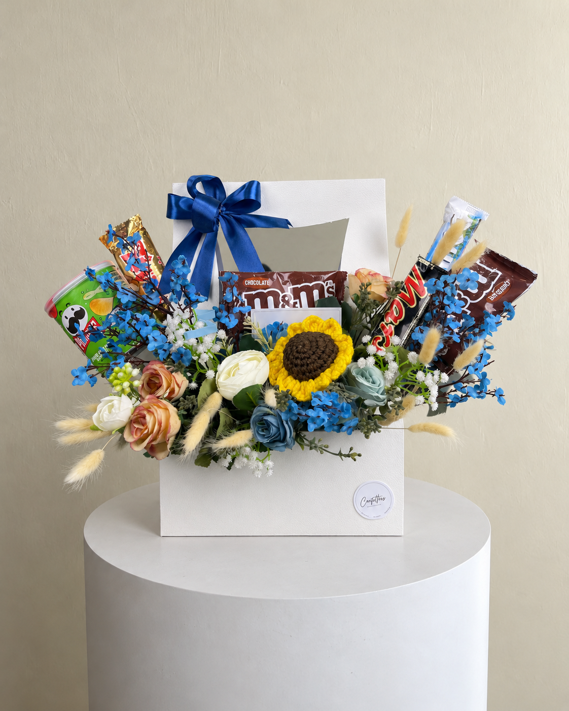 White gift box with a blue bow, chocolates, snacks, mixed floral decorations and a crochet sunflower.