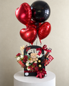 Black gift basket with a teddy bear, chocolates, red and white flowers, red heart balloons and a black balloon.