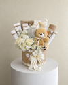 Ivory and burlap gift arrangement with a teddy bear, white flowers, wrapped gifts, a lidded glass jar and cream ribbon.
