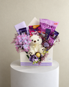 Lavender floral gift arrangement with a white teddy bear, chocolates, snacks and purple ribbon.