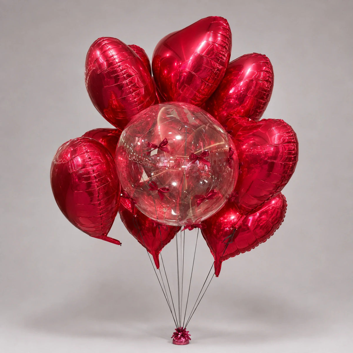 A romantic balloon bunch combining red heart-shaped foil balloons with a clear bobo balloon decorated with small burgundy bows. Coordinating ribbons and a matching balloon weight complete the arrangement for Valentine’s Day, anniversaries and romantic surprises.