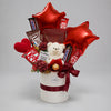 A striking red and ivory gift arrangement with a white teddy bear, two red star-shaped foil balloons, a crochet heart and assorted chocolates. KitKat, Snickers, M&M’s and a gold-wrapped chocolate sit among burgundy floral accents, presented in an ivory cylindrical gift box with burgundy ribbon.