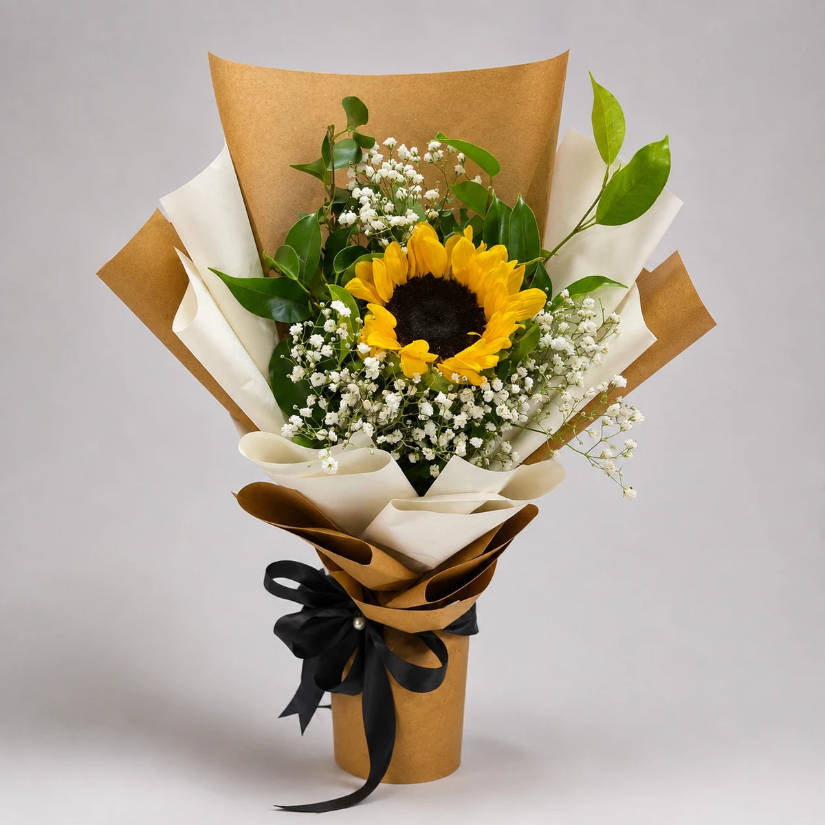 A bright sunflower takes centre stage among delicate white baby’s breath and green foliage. Kraft and ivory wrapping, tied with a black ribbon, give this cheerful bouquet a simple, warm finish for birthdays, congratulations and moments of appreciation.