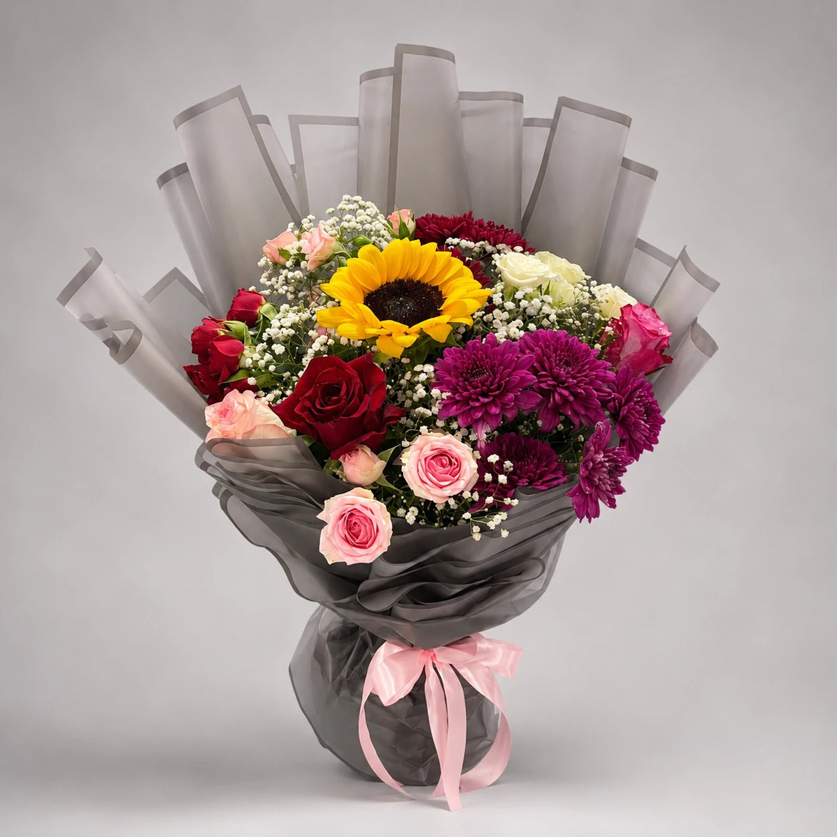 A vibrant mixed bouquet pairing a sunflower with red, pink, peach and ivory roses, purple chrysanthemums and white baby’s breath. Green foliage, layered grey wrapping and a pink ribbon complete an expressive gift for birthdays and special celebrations.