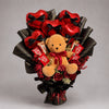 A cuddly brown teddy bear sits among red heart-shaped balloons, chocolates and deep red floral accents. Black bows and black wrapping with gold detailing add a dramatic finish to this romantic gift bouquet for Valentine’s Day, anniversaries and special birthdays.