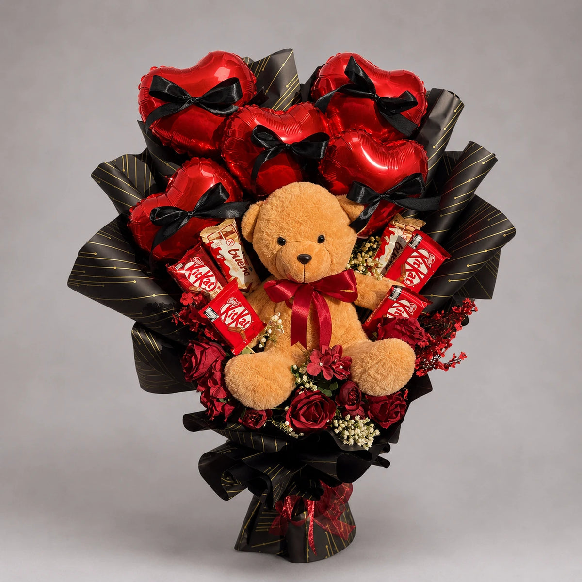 A cuddly brown teddy bear sits among red heart-shaped balloons, chocolates and deep red floral accents. Black bows and black wrapping with gold detailing add a dramatic finish to this romantic gift bouquet for Valentine’s Day, anniversaries and special birthdays.